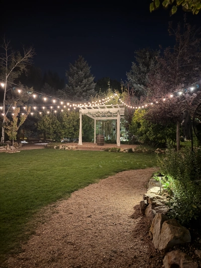 A small garden area, illuminated by string lights running from locations outside the image up to a tree above the wedding altar. There's a wooden barrel underneath the altar, and the ground is a mixture of small pebble paths and a grassy area.