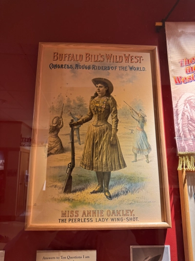 Sign from the Buffalo Bill Museum that reads "BUFFALO BILL'S WILD WEST. CONGRESS, ROUGH RIDERS OF THE WORLD. MISS ANNIE OAKLEY, THE PEERLESS LADY WING-SHOT." Annie Oakley is holding a rifle in her right hand, propped up on the prairie, with other Wild West participants taking shots in the background behind her.