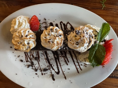 Overhead closeup of the blondie dessert from the Hungry Goat restaurant in Morrison, Colorado. Three puffs of cream are equally spaced on top of the rectangular blondie, with a chocolate cookie layer on top and sliced strawberries on opposite ends of the oval-shaped white plate. It's also drizzled with a chocolate sauce.