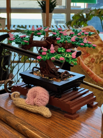 A bonsai made from Lego blocks sits on a wooden stand in a coffee shop in northwest Indianapolis. A crochet snail with a pink shell and light tan body is propped up on the side of the bonsai, and various plants are naturally lit in the blurred background.