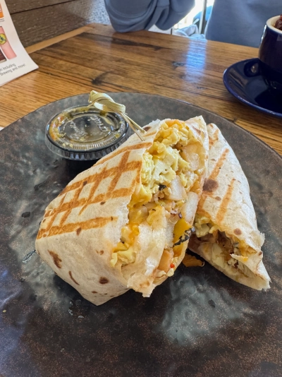 A large grilled breakfast burrito, cut in half on a diagonal and plated one half leaning on the other. It is stuffed with eggs, cheese, potatoes, onions and peppers, with a cup of somewhat spicy salsa on the site. (It was delicious every time.)