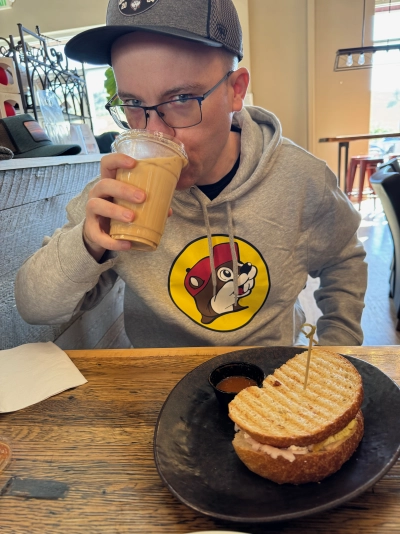 Mike Tarpey dressed in a University of Connecticut hat and a light gray Buc-ee's hoodie, drinking an iced vanilla latte and eating a turkey + egg breakfast sandwich on grilled sourdough, with a cup of vinegar-based hot sauce.