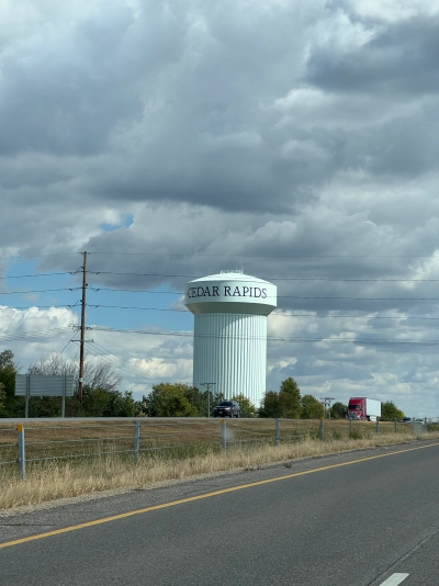 View from the driver's seat on State Road 30 eastbound in Cedar Rapids, Iowa. The water tower has the city name in big capital serif letters. There is light traffic on the westbound side of the divided highway.