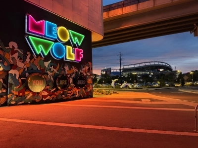 View from outside the Meow Wolf exhibit in downtown Denver, looking toward the neon "YOU ARE HERE" sign with Mile High Stadium in the background, where the Denver Broncos play home football games in the NFL. It is twilight and the sky is dark blue with an orange tint. A highway overpass is visible in the top right corner of the image.