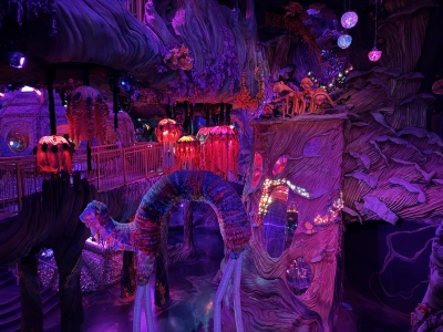 A fantastic forest landscape inside the Meow Wolf exhibit in downtown Denver. Everything is bathed in a purple light, with some orange decorative mushrooms and imaginary creatures.