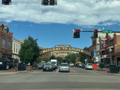 View from the driver's seat in downtown Golden, Colorado. The 12th Street stoplight is red. An arching sign reads "Howdy Folks! WELCOME TO GOLDEN, WHERE THE WEST LIVES". Small two-story buildings in earthy tones line each side of the street, with cars parked in front and pedestrians walking by.