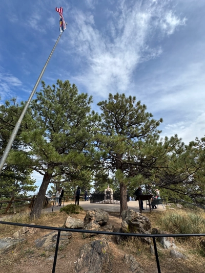 Wide-angle photograph taken from behind Buffalo Bill's grave site. The US flag and Colorado state flags fly on the same pole near the top of the image. A few tourists are observing various spots around the grave.