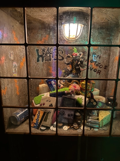 A small window exhibit inside the Meow Wolf in downtown Denver. A futuristic humanoid rat is dressed in biker clothing with a neon green mohawk. He's standing on top of a pile of retro junk, including a Nintendo Game Boy, an energy drink can, and a New York City MTA MetroCard, among many other things.