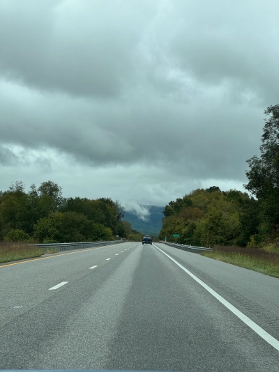 View from the driver's seat on Interstate 64 westbound near Lexington, Virginia. Trees line each side of the road, and the car in front looks like it's almost touching the low-hanging mountain clouds.