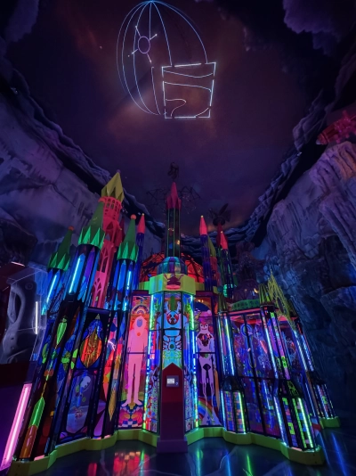 A feature exhibit of the Meow Wolf in downtown Denver. An entire miniature castle is made of stained glass in a plethora of colors. Lasers pointed upward are creating various wireframe designs on the ceiling.