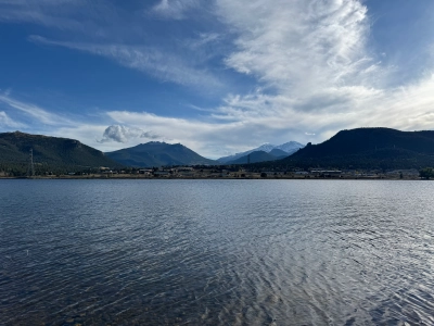 View of Lake Estes looking in the southern directions. The water is flat and hardly rippling, the sky is blue with some stretches of think white clouds, and mountains extend far into the distance.