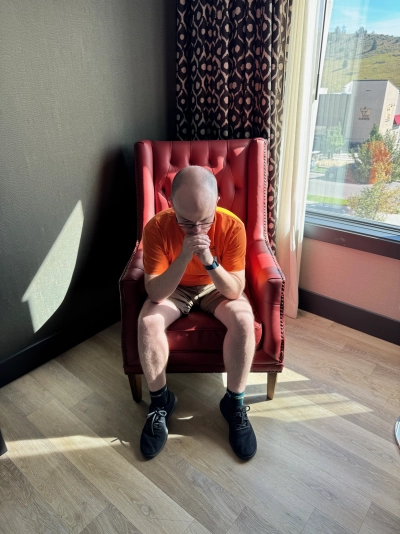 Mike Tarpey sits on a bright red chair inside the Origin Red Rocks hotel in Golden, Colorado. He is leaning forward and contemplative before the journey back home to Virginia.