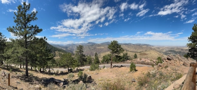 Panoramic view from the parking lot of the Buffalo Bill Museum parking lot, looking toward the Rocky Mountains underneath a partly cloudy blue sky. Trees are interspersed in the foreground, with a winding road down the mountain.