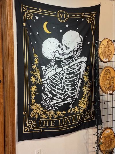 A primarily black tapestry hanging on the wall of a small indoor mall in Estes Park, Colorado. It reads "THE LOVERS" and depicts two white skeletons kissing, with arms wrapped around each other, bordered by golden flowers and the moon.