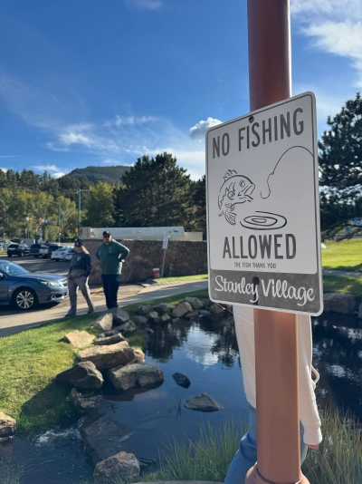 A sign that reads "NO FISHING ALLOWED. THE FISH THANK YOU. Stanley Village" mounted on a pole in front of a small pond surrounded by rocks.