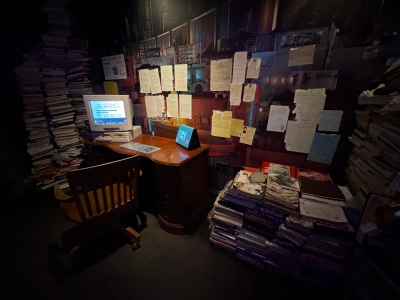 An office exhibit inside the Meow Wolf in downtown Denver. The lighting is dim, and an old Gateway computer resting on a wooden desk displays webpages from the imaginary convergence of worlds that the exhibit is based on. There are huge piles of books on either side of the desk, and letters from various fictional characters line the wall.