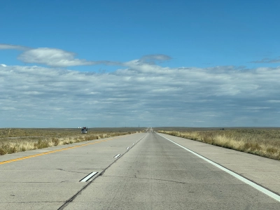 View from the driver's seat on Interstate 76 eastbound near Roggen, Colorado. The road stretches straight all the way into the cloudy horizon, with prairie grass on each side of the road.