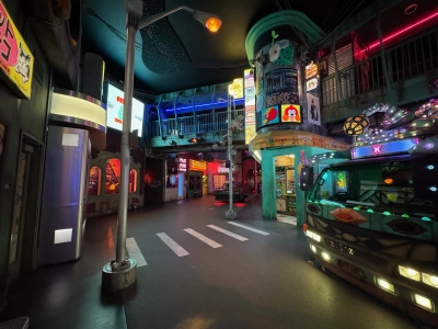 A feature exhibit of the Meow Wolf in downtown Denver. You find yourself looking at a futuristic city street, complete with fictional shops topped with neon light logos. A fake convenience store painted in teal sits behind a van on the right side, and there are lamp posts and a crosswalk.