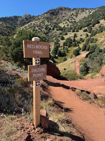 A wooden sign at the intersection of Dalton Point and Red Rocks Trail in Colorado. The red dirt trail is in the foreground and slopes steeply down. Into the distance, there's a large mountain covered by 50% trees and 50% grass. The sky is completely blue.