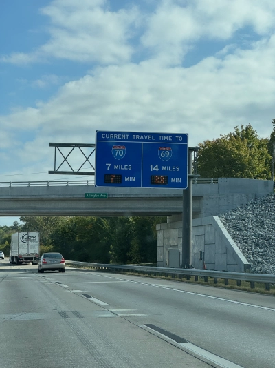 View from the driver's seat on Interstate 74 eastbound near Indianapolis. The blue highway sign reads "CURRENT TRAVEL TIME TO INTERSTATE 70, 7 MILES, 7 MINUTES. INTERSTATE 69, 14 MILES, 33 MINUTES." There is an overpass behind the sign carrying traffic on Arlington Ave.