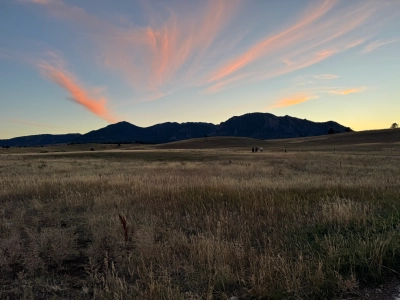 View looking west from Flatirons Vista in Colorado. There is prairie grass in the foreground, followed by some hills, and then mountains in the background. The sky is a yellowish blue at sunset, and the clouds are pinkish to orange.