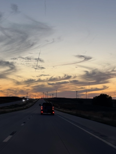 View from the driver's seat on Interstate 70 westbound near Beverly, Kansas. The sun has set and wispy clouds line the horizon. The shillouette of a wind farm is visible (about a dozen turbines).