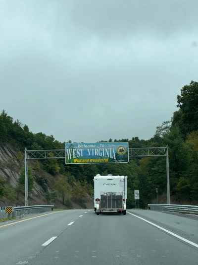 View from the driver's seat on Interstate 64 westbound at the Welcome sign for West Virginia. A small camper is underneath the sign. The speed limit sign says 70 miles per hour.