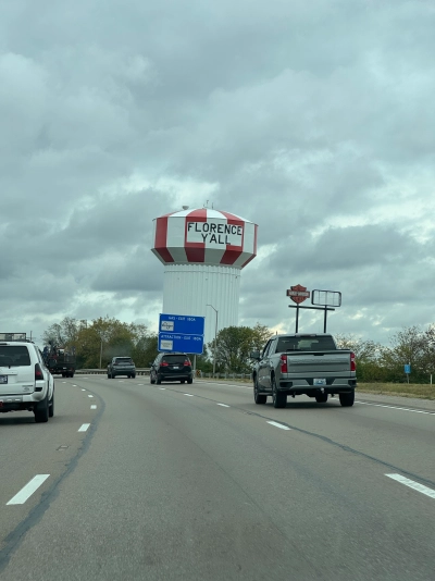 View from the driver's seat on Interstate 75 southbound near Florence, Ohio. A large red and white striped water tower reads "FLORENCE Y'ALL" under an overcast gray sky.